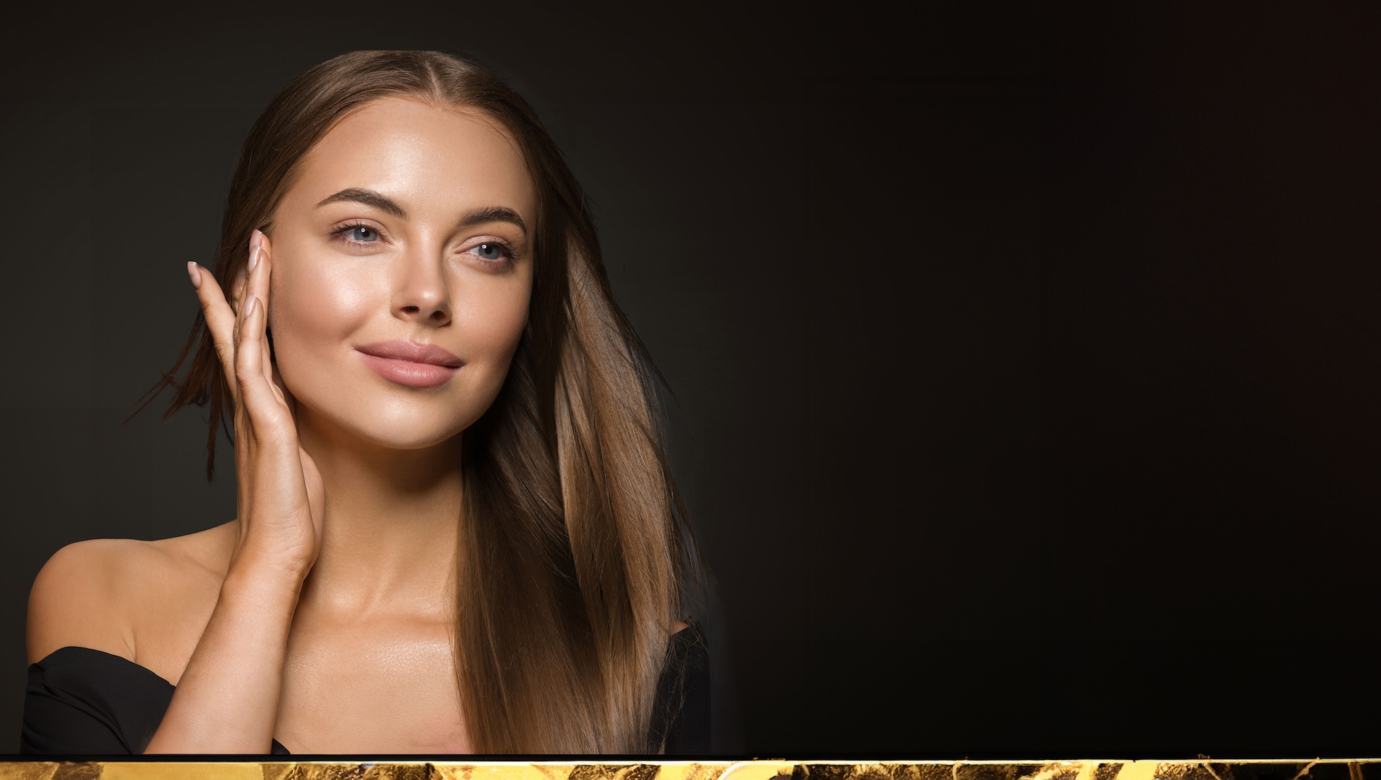 Portrait of a beautiful woman with brown hair, gently resting her hands on her face, wearing a sleek black dress against a neutral background